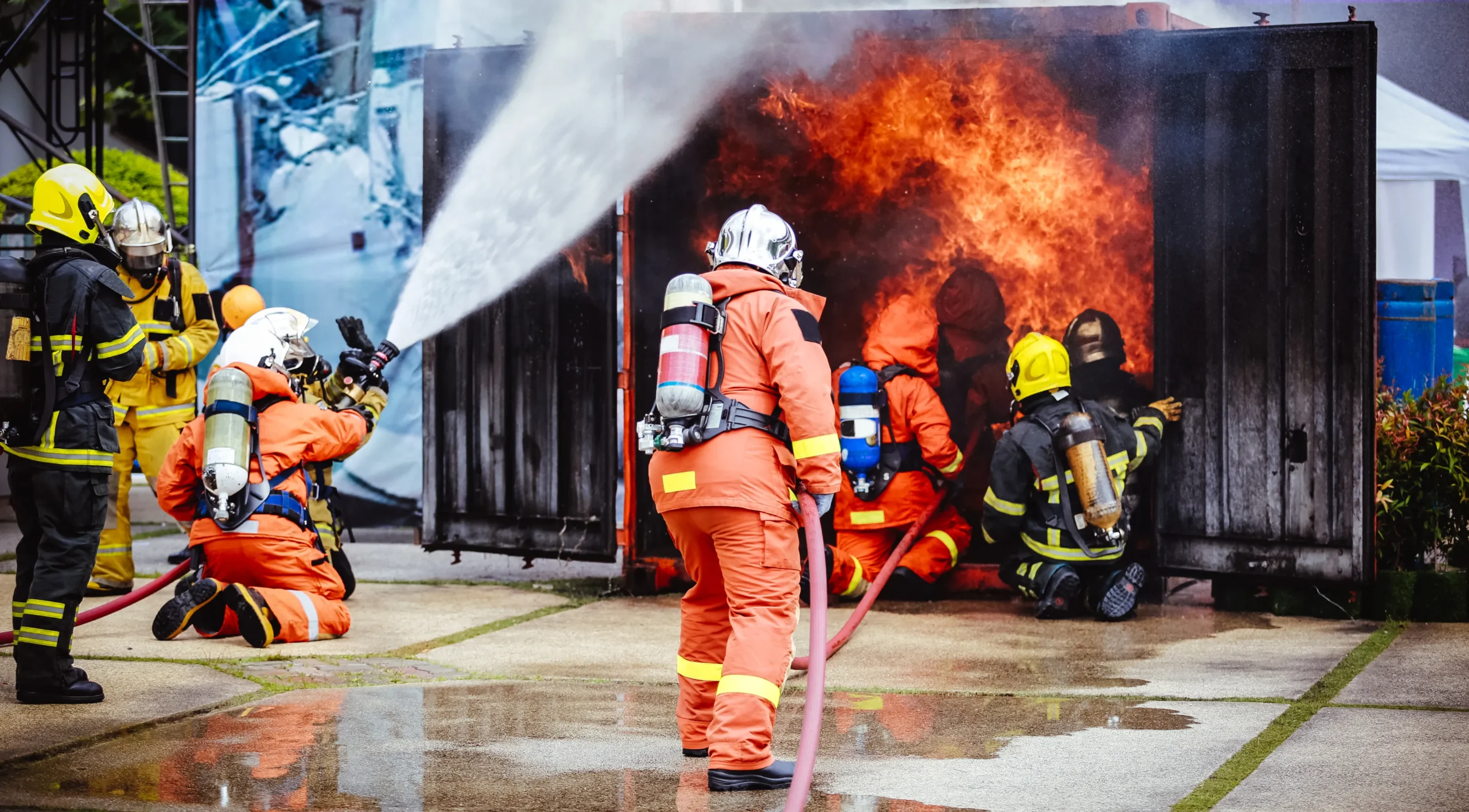 Firefighters in full gear actively extinguishing a blaze during a live fire training exercise outside a burning container.